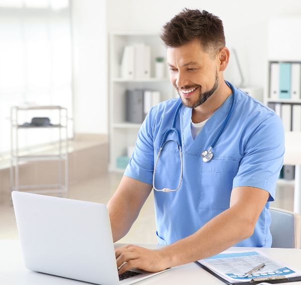 A male nurse sits at a computer and is smiling.